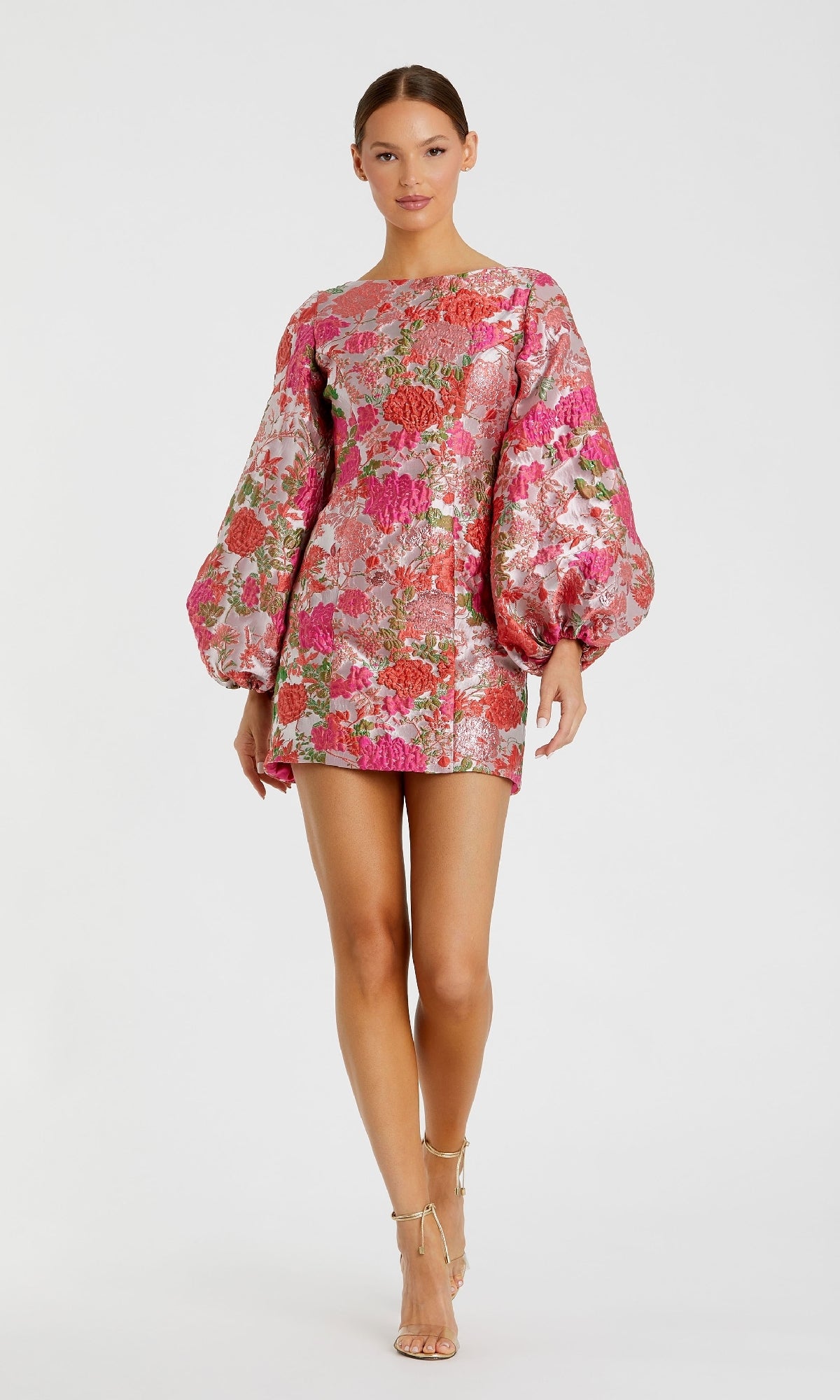 A woman models the Mac Duggal 56106 short floral wedding guest dress with voluminous sleeves and metallic pink, red, and green patterns, styled elegantly as a cocktail dress against a plain white background.